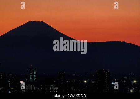 Schwarze Silhouette des Vulkankegels des fuji, der vor einem leuchtend orangen Sonnenuntergang mit den Lichtern der Stadt tokio im Vordergrund steht Stockfoto