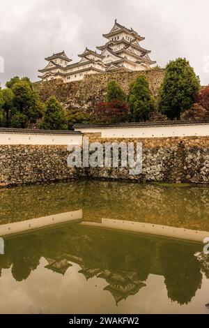 Die Burg himeji über einer befestigten Mauer spiegelt sich im Wasser eines Spiegelteichs unter einem dunklen Himmel, der einen Regensturm bedroht Stockfoto