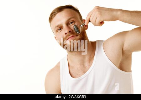 Junger Mann pflegt seine Gesichtshaut, entfernt Schwellungen mit Massagewalze vor weißem Hintergrund. Kopierbereich. Anzeige Stockfoto