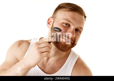 Der junge Mann pflegt seine Gesichtshaut, entfernt Alterserscheinungen und strafft seine Gesichtskonturen mit einem Massagewalzen vor weißem Hintergrund. Stockfoto