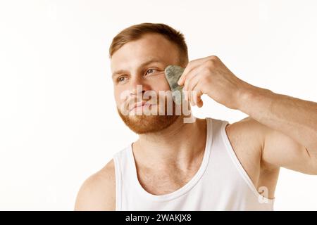 Porträt eines jungen Mannes im T-Shirt, der seine Gesichtshaut mit Massageabstreifer vor weißem Hintergrund pflegt. Konzept der Anti-Aging-Verfahren. Stockfoto