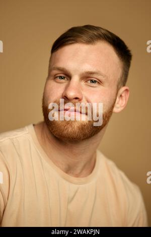 Porträt eines jungen hübschen rothaarigen Mannes mit lächelndem Bart, der die Kamera vor sandigem Farb-Studio-Hintergrund betrachtet. Stockfoto