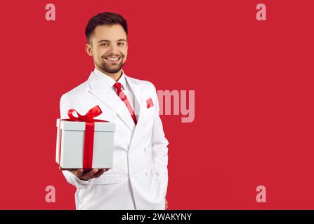 Glücklicher, großzügiger junger Mann im Anzug, der eine Geschenkbox hält und auf rotem Hintergrund lächelt. Stockfoto