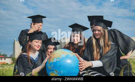 Absolventen in schwarzen Gewändern untersuchen einen geografischen Globus, der auf dem Gras sitzt. Stockfoto
