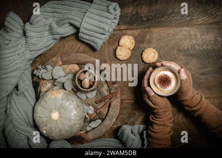 Gemütliche Kaffeeszene: Weibliche Hand hält eine Tasse Cappuccino, Holzteller mit Kürbis und Herbstdekorationen, grüner Wollpullover, hölzerner Hintergrund, Oberseite Stockfoto