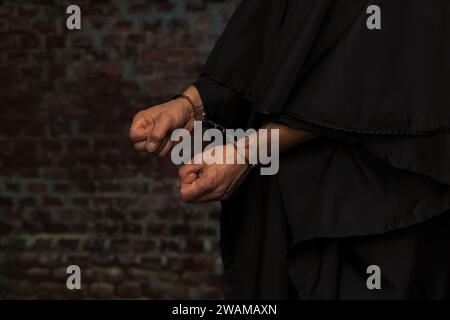 Die Hände eines Mönchs oder Priesters in Handschellen auf dem Hintergrund einer Steinmauer. Begriff: Alte Straftat, strafrechtliche Haftung, religiöse Figur. Stockfoto