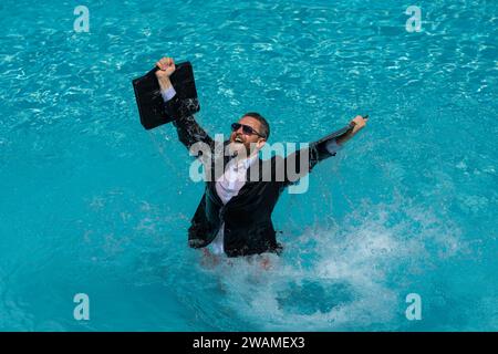 Ein lustiger, überraschter Geschäftsmann im Anzug mit einem Laptop im Swimmingpool. Verrückter, aufgeregter Geschäftsmann im Sommerurlaub. Aufgeregter Geschäftsmann in Neoprenanzug aufgeregt Stockfoto