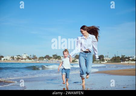 Authentisches Porträt der kaukasischen glücklichen Mutter und Tochter lächeln, während sie entlang der Atlantikküste laufen und dabei wunderschöne Wellen genießen, die sich während der pou die Füße waschen Stockfoto