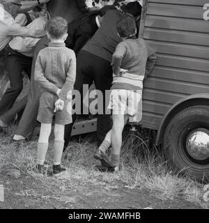 1970er Jahre, historisch, Sommer und draußen auf einem Feld beobachten zwei Jungen, wie eine Reihe erwachsener Männer versuchen, ein Pferd in seinen Radwagen oder Pferdekasten zu schieben, auf einer Dampfmesse in England, Großbritannien. Stockfoto