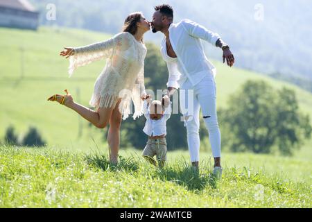 Multiethnische Eltern und gemischtes Baby, das auf Sommergras läuft. Multiethnisches Paar küsst die Natur. Birassisches multiethnisches Baby-Kind erster Schritt Stockfoto