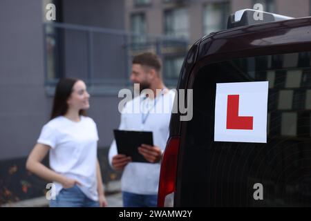 Fahrer und Kursleiter mit Klemmbrett in der Nähe des Autos im Freien, selektiver Fokus auf L-Platte. Fahrschule Stockfoto