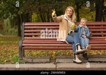 Glückliche Mutter und ihr Sohn verbringen Zeit zusammen mit trockenen Blättern auf Holzbank im Herbstpark Stockfoto