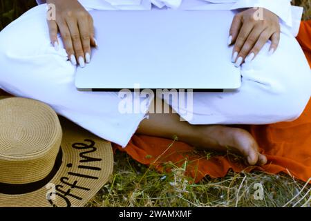 Mädchen in weißer Luxusbekleidung sitzt im Sommer auf grünem Gras mit Laptop auf den Knien. Remote-Job, Online-Arbeit. Strohhut mit Text OFFLINE. Reiche Frau. Stockfoto