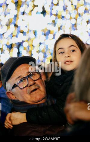 Latina, Italien. Januar 2024. Die Weihnachtslichter bilden die Kulisse für einen Großvater und seine Enkelin, die faszinierend den Abstieg der Flying Befana anlässlich der Epiphany Feiern in Latina beobachten. Hunderte jubelnde Kinder und Erwachsene begrüßten die Befana, die anlässlich der Epiphanienfeier in Latina vom Turm des Rathauses abstieg. (Bild: © Marcello V Credit: ZUMA Press, Inc./Alamy Live News Stockfoto