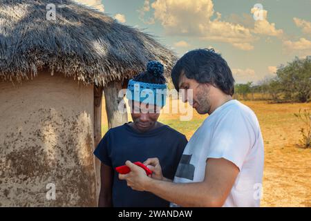 Porträt eines afrikanischen Dorfmannes, der auf ein Telefon schaut und mit einem NGO-Mitarbeiter diskutiert, vor der Hütte bei Sonnenuntergang, Dorf in Botswana Stockfoto