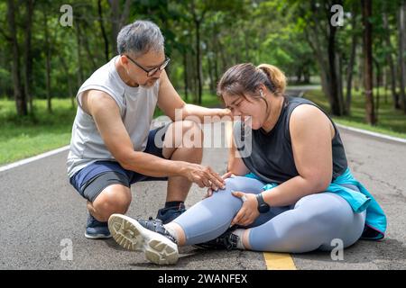 Übergewichtige junge Frau mit Knieschmerzen, die auf der Laufbahn im örtlichen Park sitzt und beide Hände am Knie greifen, während ein Mann mittleren Alters k Stockfoto