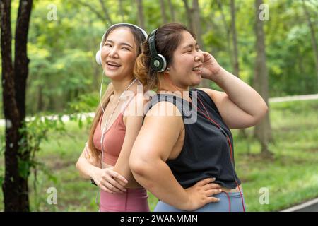 Zwei glückliche asiatische Freundinnen, eine übergroße und eine schlanke, stehen mit dem Rücken zusammen und hören Musik von ihren Kopfhörern in einem lokalen Park Stockfoto