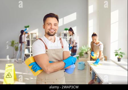 Glücklicher männlicher junger Hausmeister in Uniform, der in die Kamera schaut, die Haushaltsreinigungsbedarf hält Stockfoto