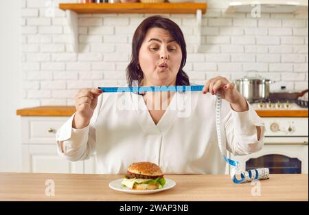 Eine fette, übergewichtige junge Frau, die in der Küche sitzt und ein Maßband mit einem Burger auf dem Tisch hält. Stockfoto