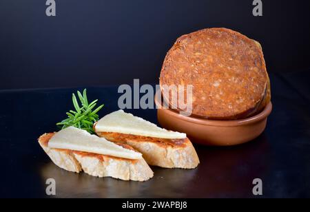 Spanische traditionelle Tapas von Sobrassada, die auf gerösteten Brotgewürzen aufgetragen werden, serviert mit Kurkäse und einer Tonschale auf dunklem Hintergrund. Stockfoto