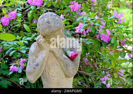 Die Trauernde (Steinfigur) mit Blume vor Rhododendronbüsche Stockfoto