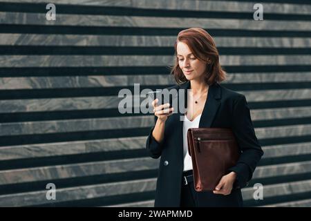 Lächelnde Geschäftsfrau mit Smartphone und Ledermappe im Freien. Frau mittleren Alters mit Ingwerhaartippung auf Smartphone. Stockfoto