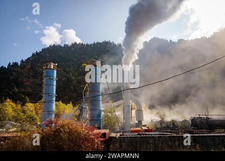 Industrielandschaft mit rauchenden Fabrikschornsteinen vor dem Hintergrund bergigen Geländes, die den Einfluss menschlicher Aktivitäten auf die Umwelt widerspiegelt Stockfoto