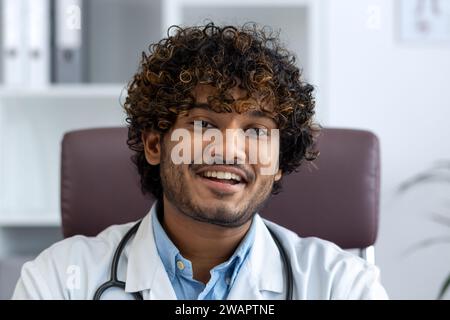 Close-up Porträt des Arztes in der Klinik am Arbeitsplatz, Mann in weißem medizinischen Mantel lächelnd in die Kamera, zufriedener indischer Mann mit lockigen Haaren. Stockfoto