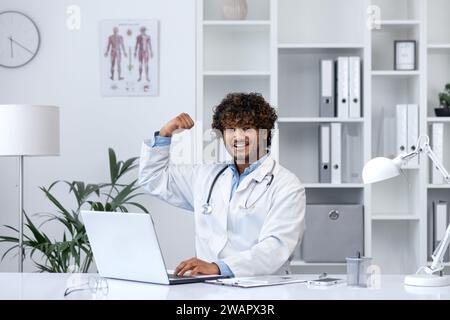 Gut gemacht Superheldenarzt, Mann lächelt in die Kamera, hält seine Hände hoch, freut sich über den Erfolg bei der Behandlung des Patienten, arbeitet in der Klinik in der medizinischen Praxis, sitzt am Tisch. Stockfoto