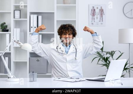 Gut gemacht Superheldenarzt, Mann lächelt in die Kamera, hält seine Hände hoch, freut sich über den Erfolg bei der Behandlung des Patienten, arbeitet in der Klinik in der medizinischen Praxis, sitzt am Tisch. Stockfoto