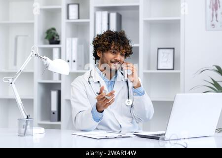 Junger erfolgreicher hispanischer Arzt in der Klinik im medizinischen Büro kommuniziert freudig am Telefon, der Klinikmitarbeiter sitzt am Schreibtisch und arbeitet mit einem Laptop, berät Patienten. Stockfoto