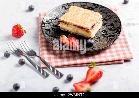 Ein Stück Napoleonkuchen mit Mohnsamen und Kirschen, dekoriert mit Erdbeeren und Blaubeeren auf hellem Hintergrund. Schließen Stockfoto