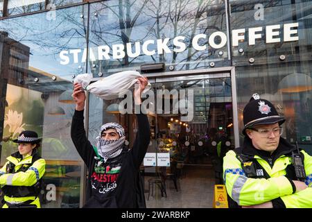 Manchester, Großbritannien. Januar 2024. Ein Demonstrant in Manchester, Großbritannien, hält ein vermeintliches totes Kind in einem Grabtuch vor dem Starbucks Coffee Deansgate. Palästinensische Proteste im Zentrum von Manchester. UK. Über zweitausend Demonstranten versammelten sich auf dem Petersplatz und forderten einen Waffenstillstand. Sie marschierten dann durch das Stadtzentrum. Die Polizei bewachte Filialen, von denen die Demonstranten sagten, dass sie Verbindungen zu Israel hatten, einschließlich Barclays Bank und Starbucks Coffee. Die Demonstranten schwenkten Fahnen und trugen Plakate mit Slogans gegen den Krieg. Manchester UK. Bild: Garyroberts/worldwidefeatures.com Kredit: GaryRobertsphotogra Stockfoto