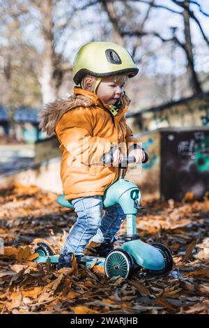 Entzückender Kleinkind mit gelbem Schutzhelm, der am Herbsttag im Freien auf dem Roller reitet. Kindertraining auf dem Minifahrrad im Stadtpark. Lustige Herbstaktivitäten im Freien für kleine Kinder Stockfoto