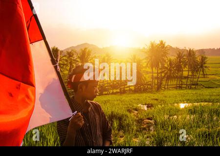 asiatischer Teenager in traditioneller javanischer Kleidung, der die indonesische Flagge selbstbewusst mit optischem Flair zur Seite hält Stockfoto