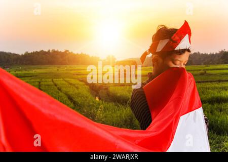 Asiatischer Teenager in traditioneller javanischer Kleidung mit indonesischer Flagge und Küssen mit Weisheit auf natürlichem Hintergrund in grünen Reisfeldern Stockfoto