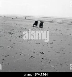 1960er Jahre, historisch, an einem weiten, leeren Strand, zwei Personen sitzen auf Liegestühlen und genießen die Sonne, England, Großbritannien. Stockfoto