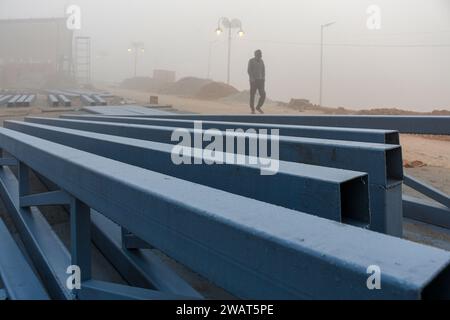 Ein Mann läuft an einem nebeligen Morgen vor dem Besuch des indischen Premierministers Narendra Modi in Ayodhya an Baumaterialien entlang, die auf einem neu gebauten Bürgersteig gelegt wurden. Ramnagari Ayodhya, einst bekannt für seine engen Gassen und verfallenen Straßen, macht stetige Fortschritte: Alle Straßen und Gebäude, die zum RAM Janmabhoomi führen, werden erweitert und renoviert, um ein reibungsloses Reisen zu ermöglichen. Stockfoto