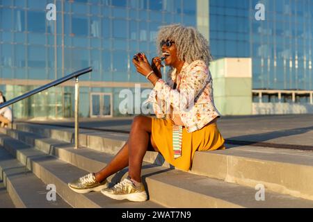 Ausdrucksstarke, schwarze Dame mit grauem Afro-Haar und Sonnenbrille auf blauem Lippenstift im sonnigen Stadtzentrum. Konzept: pro-Aging, Selbstvertrauen Stockfoto