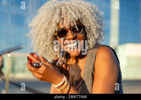 Ausdrucksstarke, schwarze Dame mit grauem Afro-Haar und Sonnenbrille auf blauem Lippenstift im sonnigen Stadtzentrum. Konzept: pro-Aging, Selbstvertrauen Stockfoto