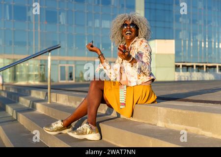 Ausdrucksstarke, schwarze Dame mit grauem Afro-Haar und Sonnenbrille auf blauem Lippenstift im sonnigen Stadtzentrum. Konzept: pro-Aging, Selbstvertrauen Stockfoto