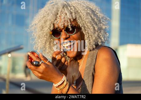 Ausdrucksstarke, schwarze Dame mit grauem Afro-Haar und Sonnenbrille auf blauem Lippenstift im sonnigen Stadtzentrum. Konzept: pro-Aging, Selbstvertrauen Stockfoto