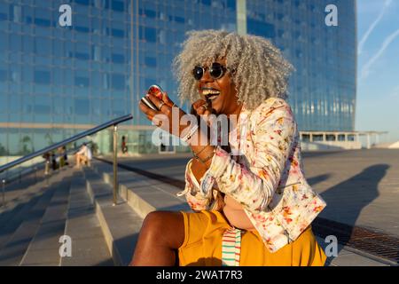 Ausdrucksstarke, schwarze Dame mit grauem Afro-Haar und Sonnenbrille auf blauem Lippenstift im sonnigen Stadtzentrum. Konzept: pro-Aging, Selbstvertrauen Stockfoto