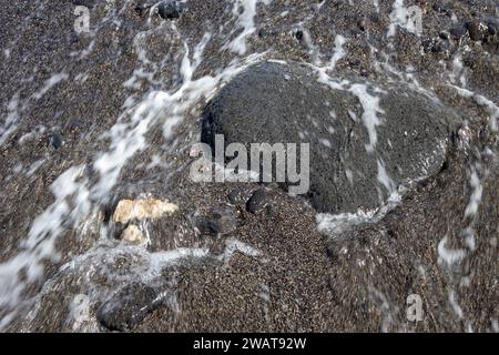 Dunkler vulkanischer Stein, vom Wasser des Atlantiks am Strand gewaschen. Wasser in Bewegung. Lanzarote, Kanarische Inseln, Spanien. Stockfoto