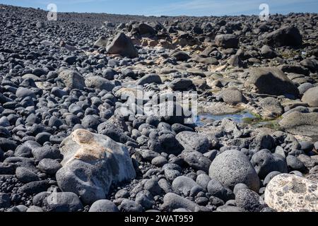 Dunkle vulkanische Steine an der Küste des Atlantischen Ozeans. Blauer Himmel im Hintergrund. Kleines Paddel. Lanzarote, Kanarische Inseln, Spanien. Stockfoto