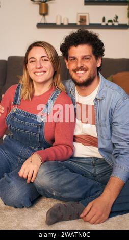 Zwei kaukasische fröhliche junge Erwachsene, die verliebt in die lächelnde Kamera schauen, die auf Teppich im Wohnzimmer sitzt. Stockfoto