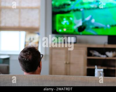 Kopf eines Mannes, der Videospiele spielt, die von hinten gesehen werden, während er auf einer Couch mit TV-Monitor sitzt und ein Flugzeug-Jet-Kampfflugzeug-Videospiel auf den Bildschirm projiziert. Stockfoto