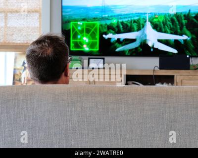 Kopf eines Mannes, der Videospiele spielt, die von hinten gesehen werden, während er auf einer Couch mit TV-Monitor sitzt und ein Flugzeug-Jet-Kampfflugzeug-Videospiel auf den Bildschirm projiziert. Stockfoto