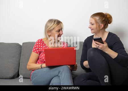 Zwei Frauen sitzen zu Hause auf der Couch und unterhalten sich miteinander und kaufen online per Smartphone und Laptop. Stockfoto