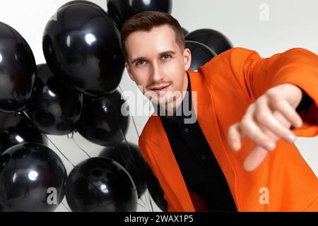 Schöner Mann in orangefarbener Jacke mit schwarzen Luftballons auf weißem Hintergrund. Geburtstagsfeier Stockfoto